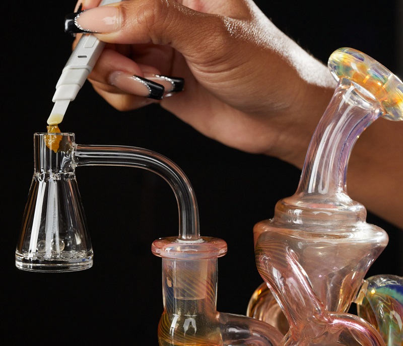 A glass dab rig with quartz banger and a female hand using a hot knife dab tool to drop some wax into the dab rig